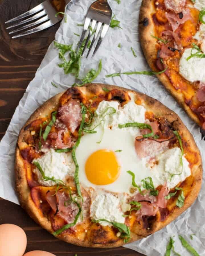 two ham and cheese breakfast pizzas on a dark wood backdrop with forks and eggs on the sides as garnishes.
