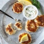 Four mini crab cakes on a silver metal plate with another crab cake cut in half. Lemon garnish and a bowl of sauce.