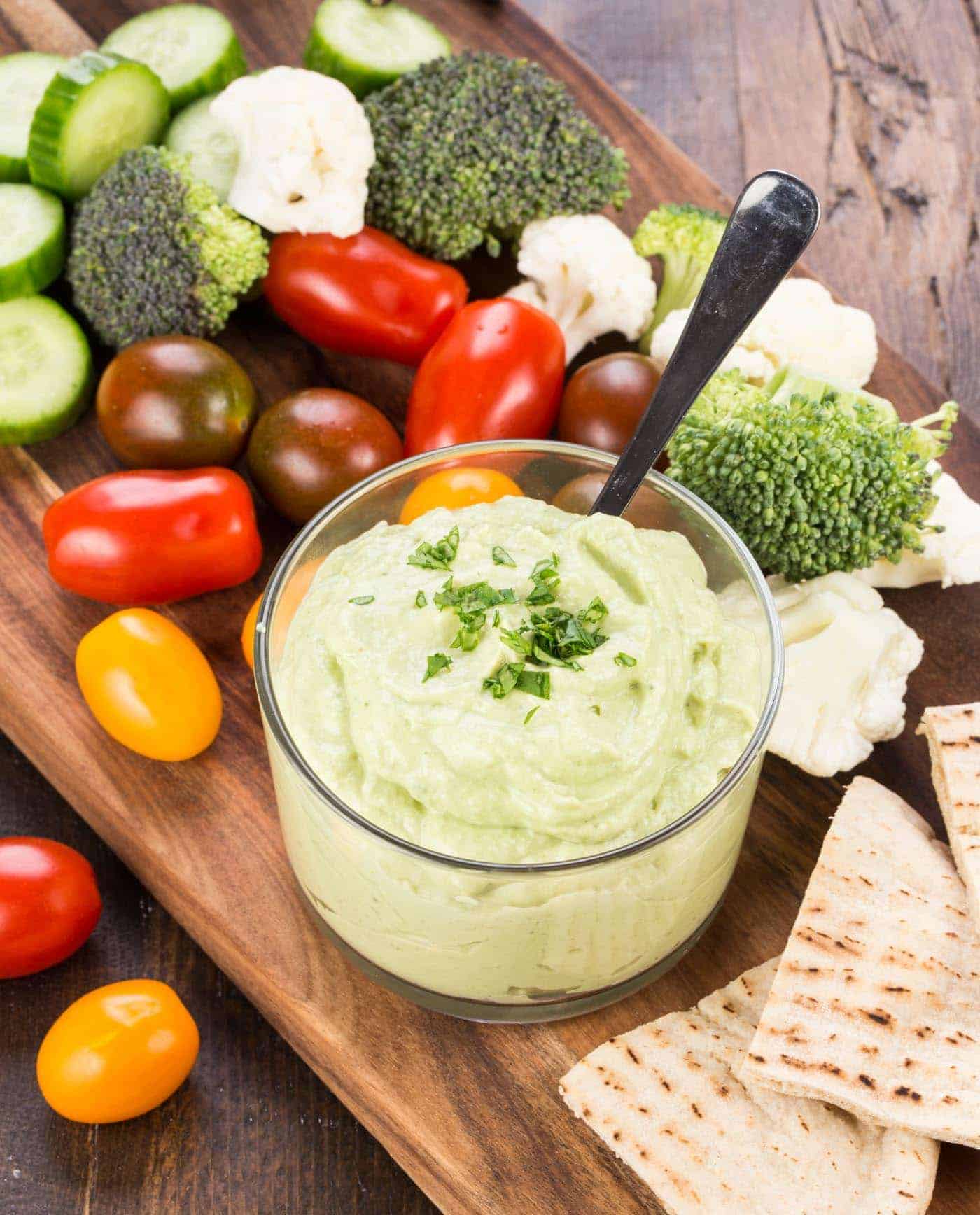 bowl of  Dip on wooden board surrounded by colorful vegetables and pita wedges.