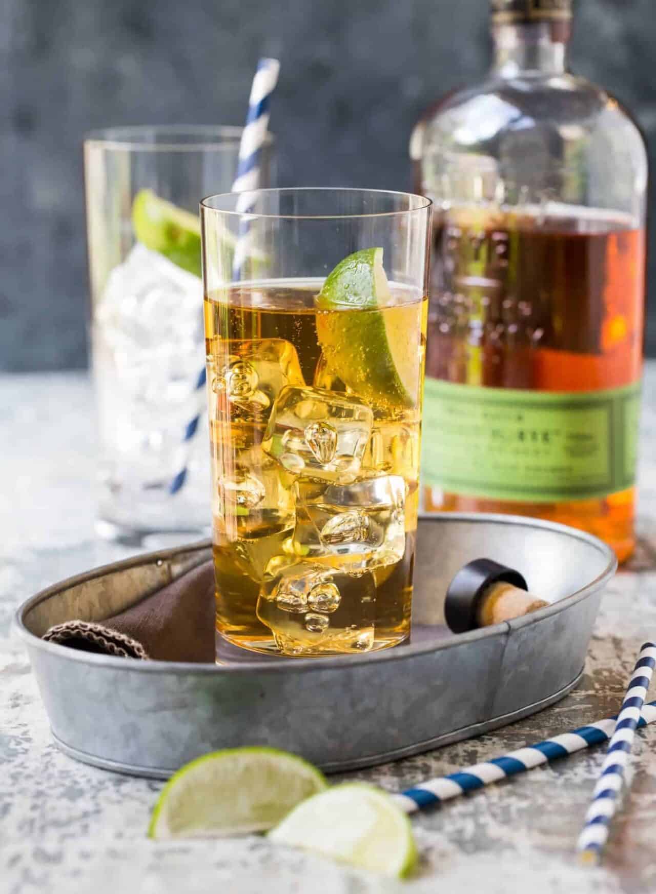 A glass of bourbon and ginger next to limes, straws, and a bottle of bourbon. 