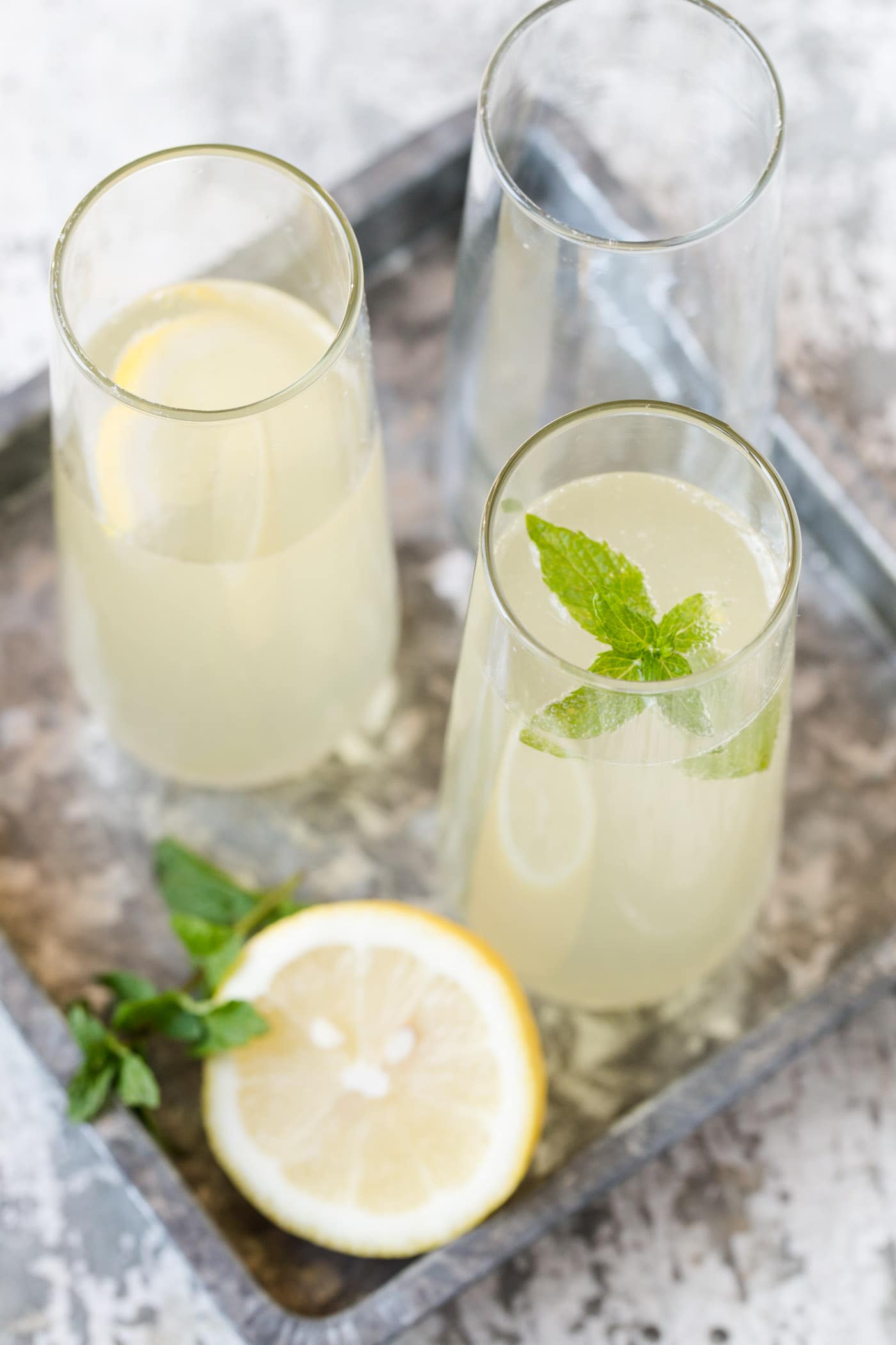 Cocktails in stemless champagne flutes on a mottled silver tray with mint and a lemon wedge for garnish.