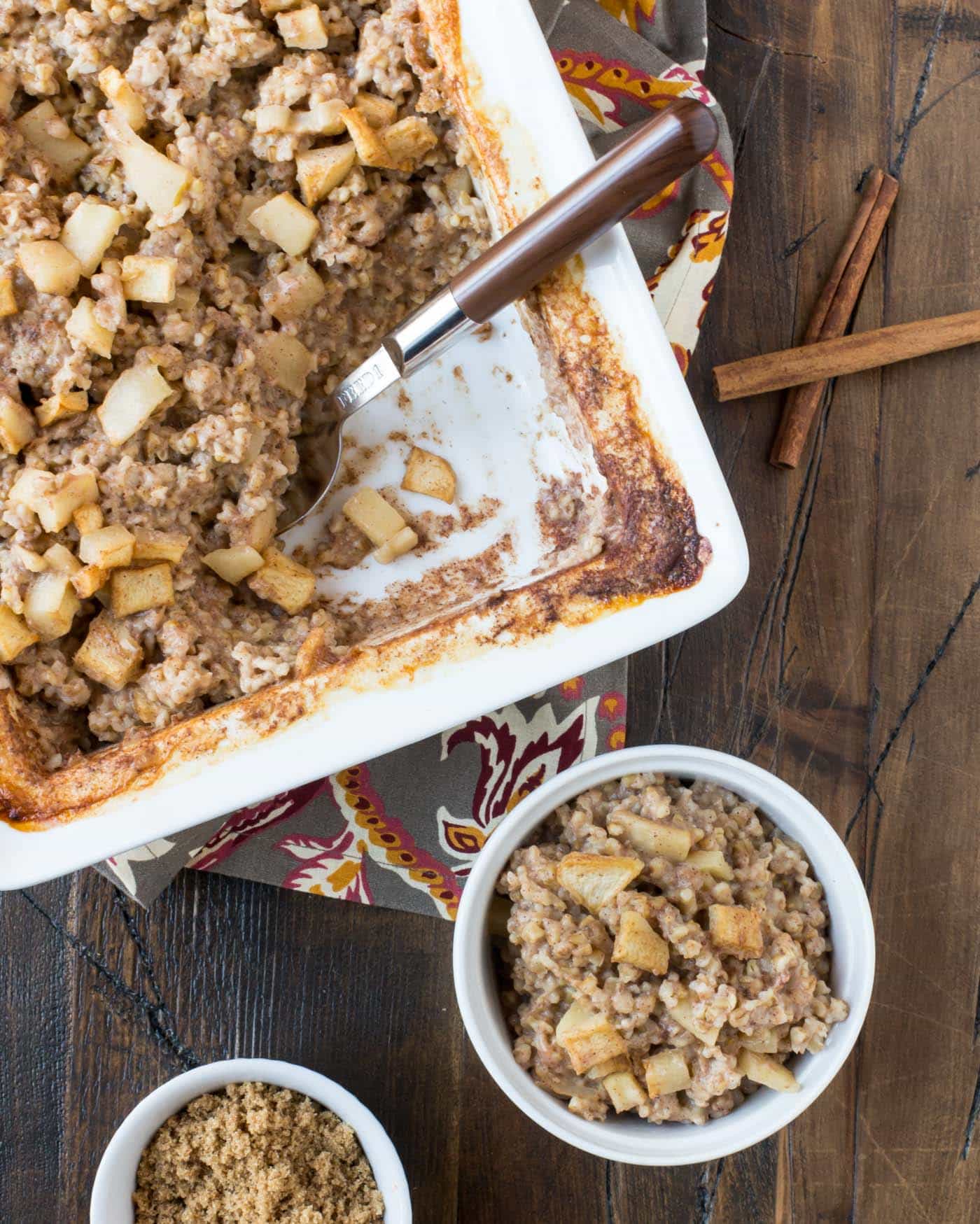 Top down image of white pan filled with baked oatmeal and a brown spoon for serving with a cinnamon stick for garnish.