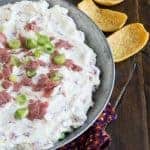 chipped beef dip in a metal baking dish on a dark background with corn chips for eating.