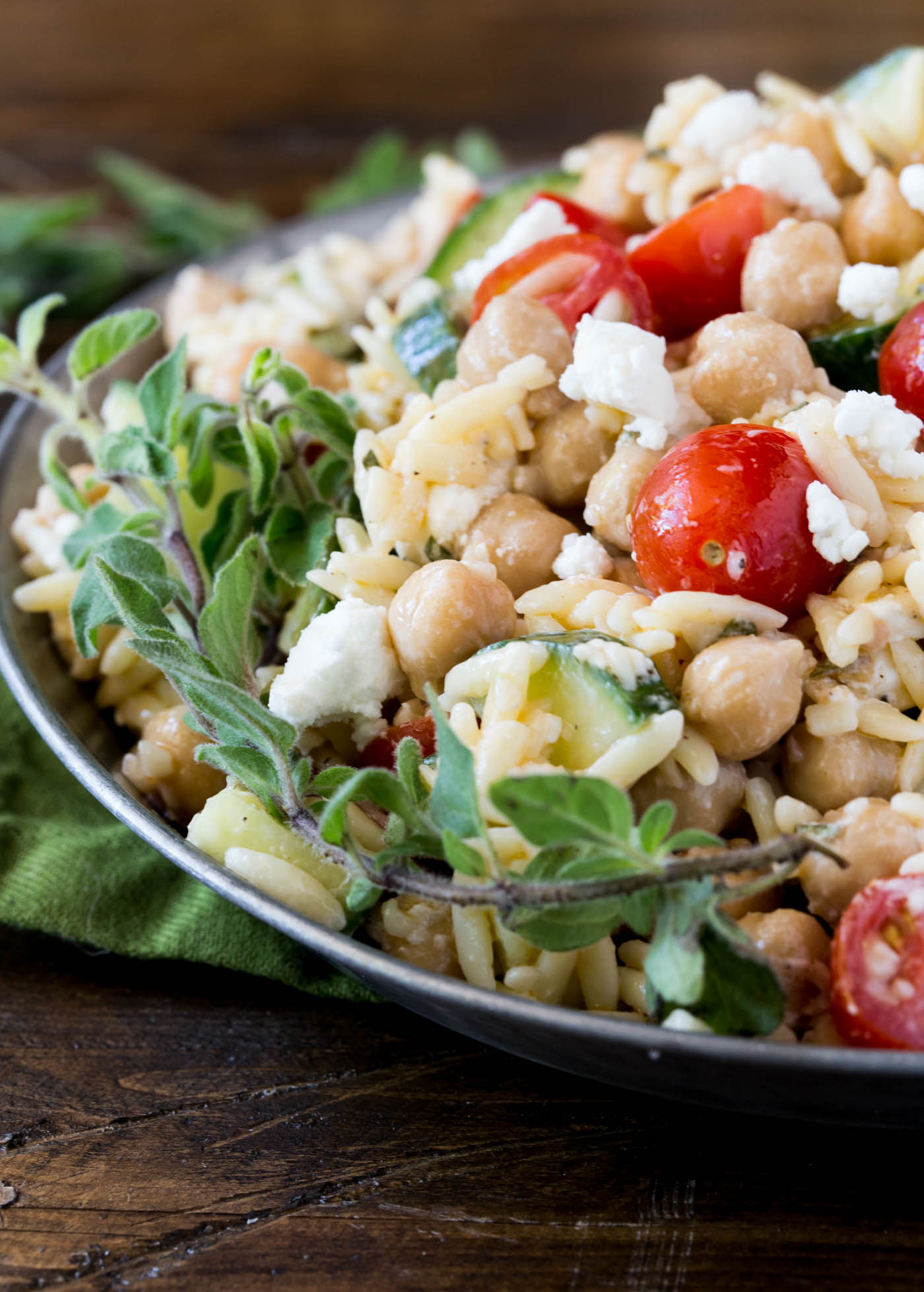 Silver bowl filled with orzo and chickpea salad. Garnished with fresh oregano, tomatoes and cucumbers. 