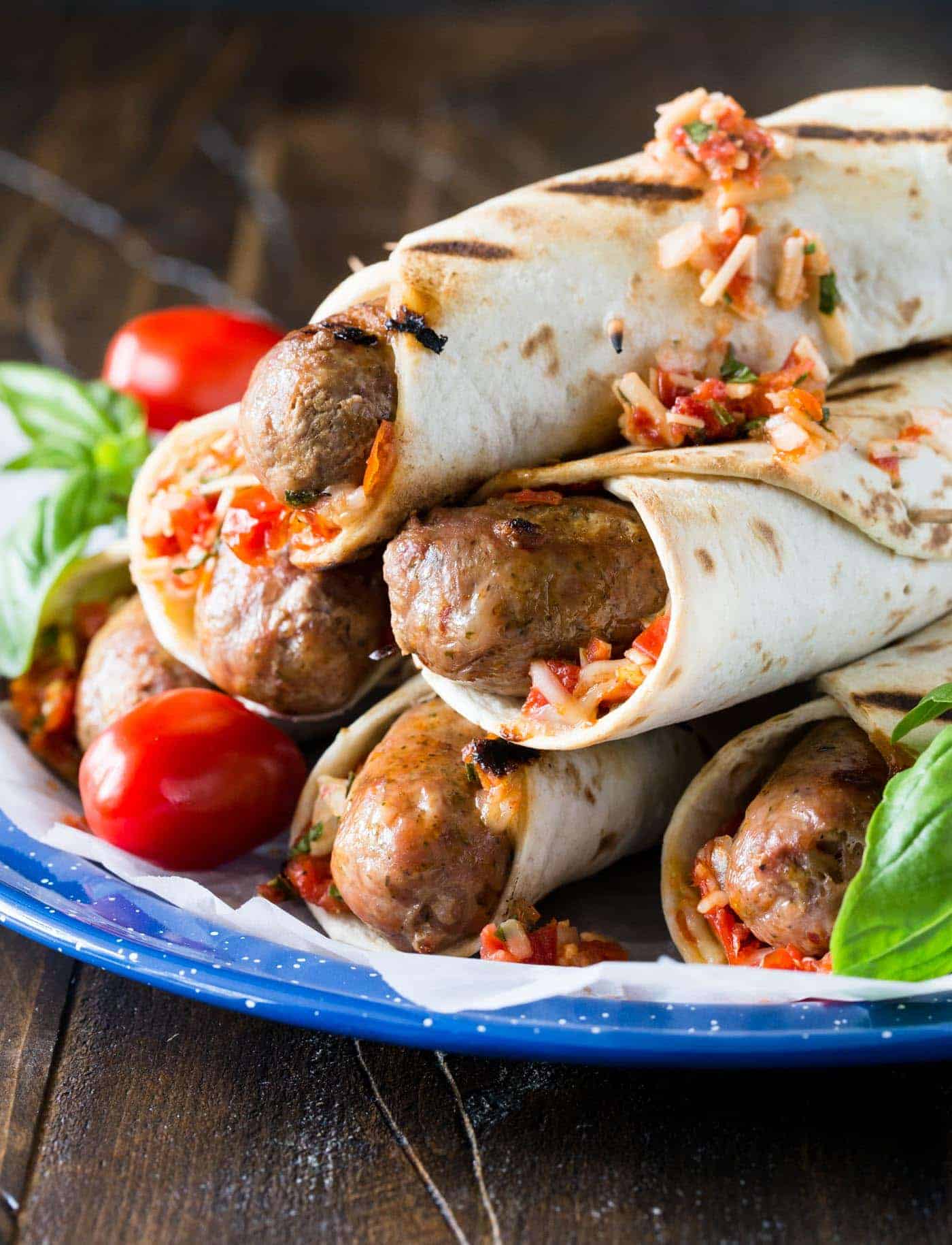 Grilled Italian Sausages on a blue plate. Sausages are wrapped in grilled flatbreads and garnished with a roasted tomato, asiago cheese and basil relish.