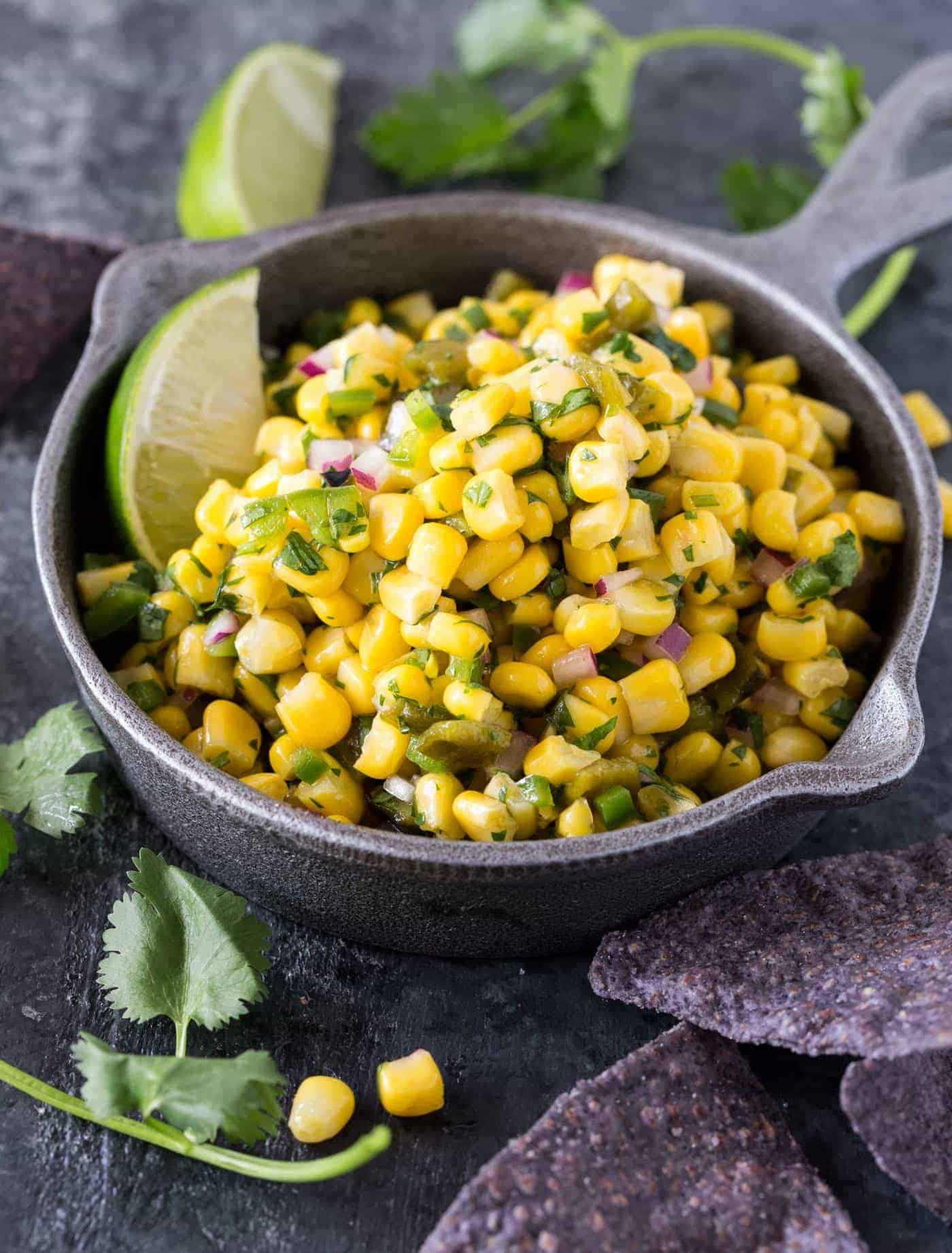 Bowl of Corn Salsa recipe surrounded by chips, cilantro and lime wedges. 