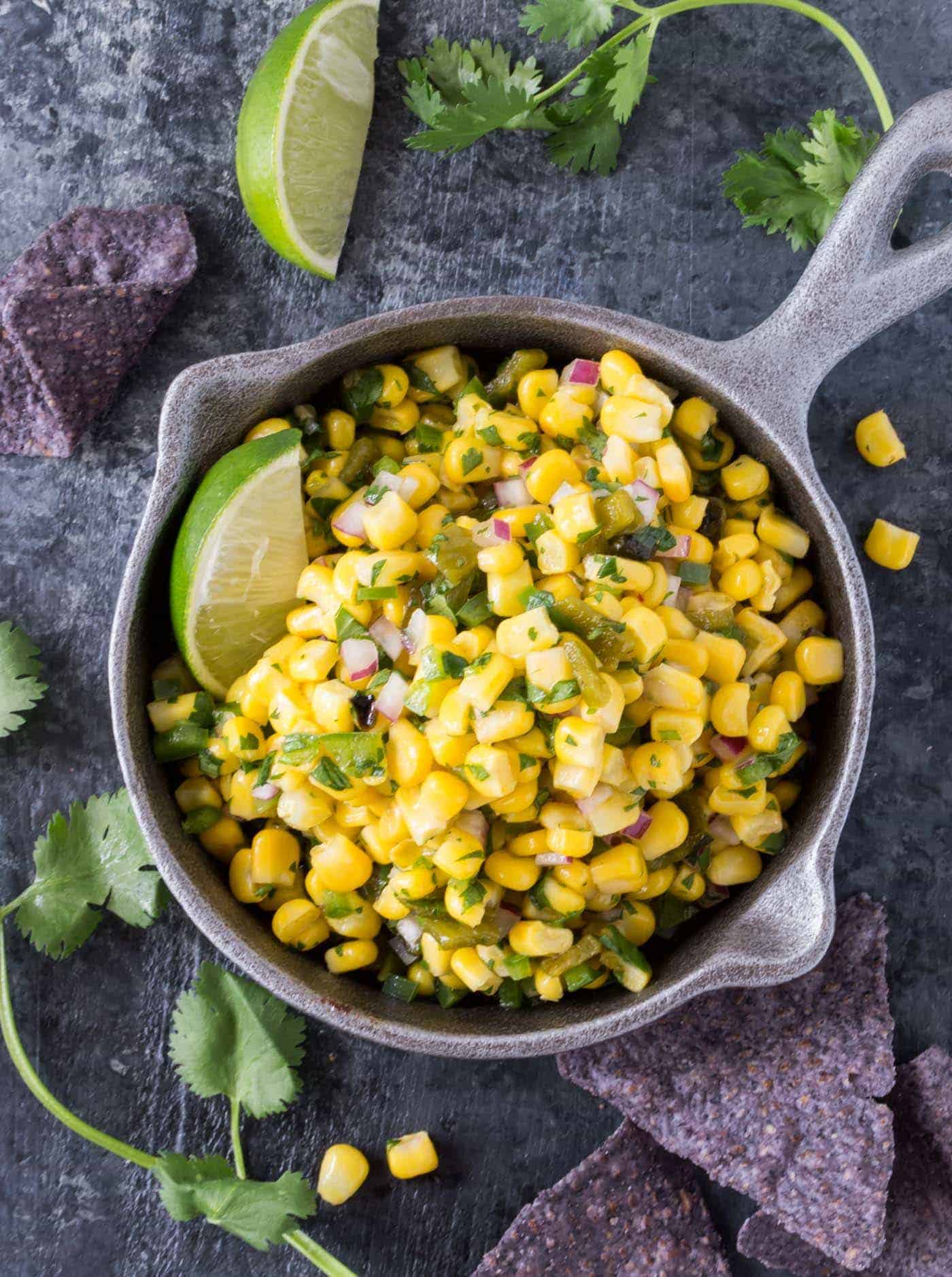 Bowl of Copycat Chipotle Corn Salsa recipe surrounded by chips, cilantro and lime wedges. 