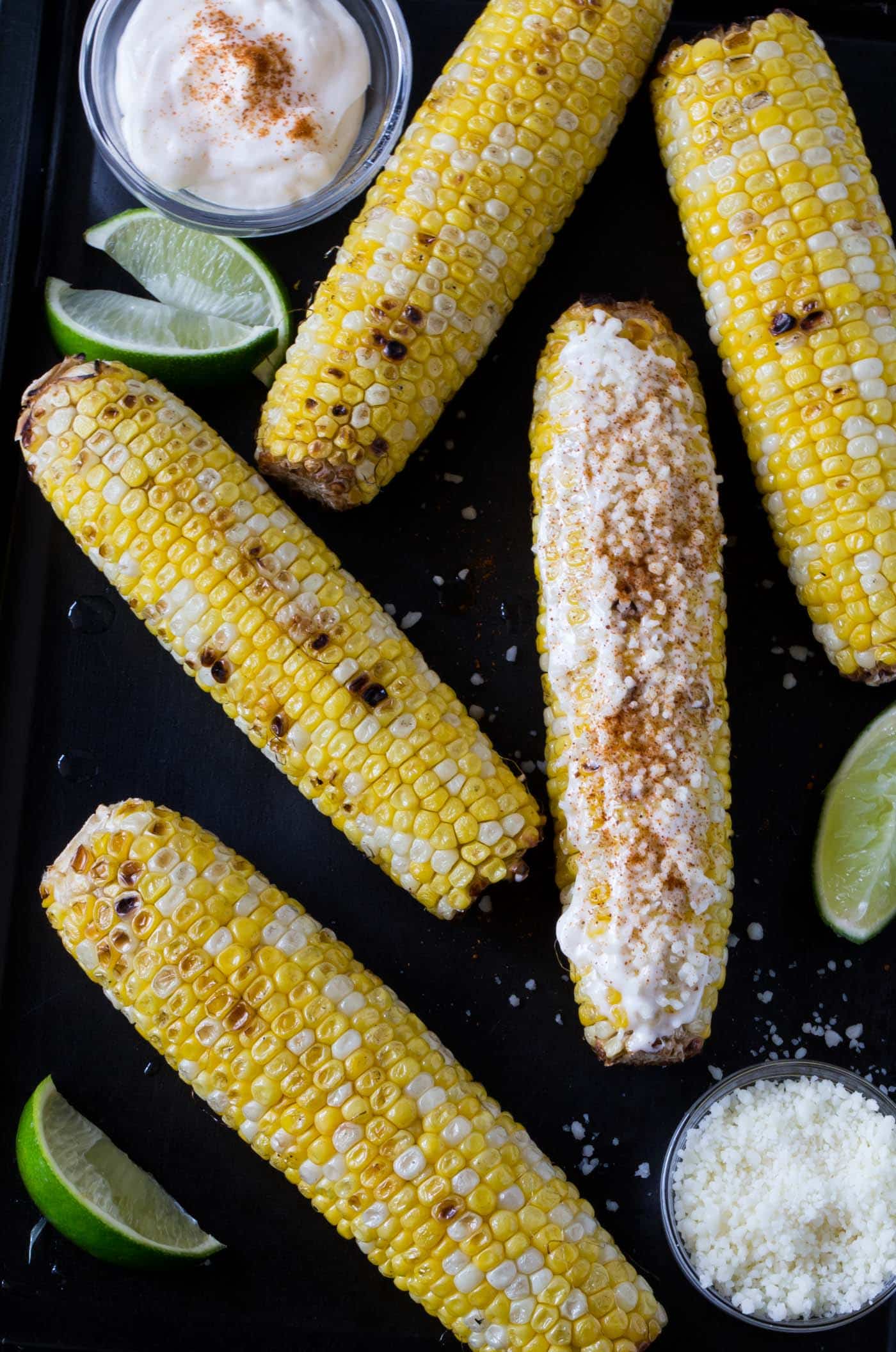 Mexican Grilled Corn with mayonnaise, lime wedges, cotija cheese and cayenne pepper on a sheet pan