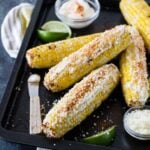 Mexican grilled corn with mayonnaise, cotija cheese, cayenne pepper and lime wedges piled high on a sheet pan.