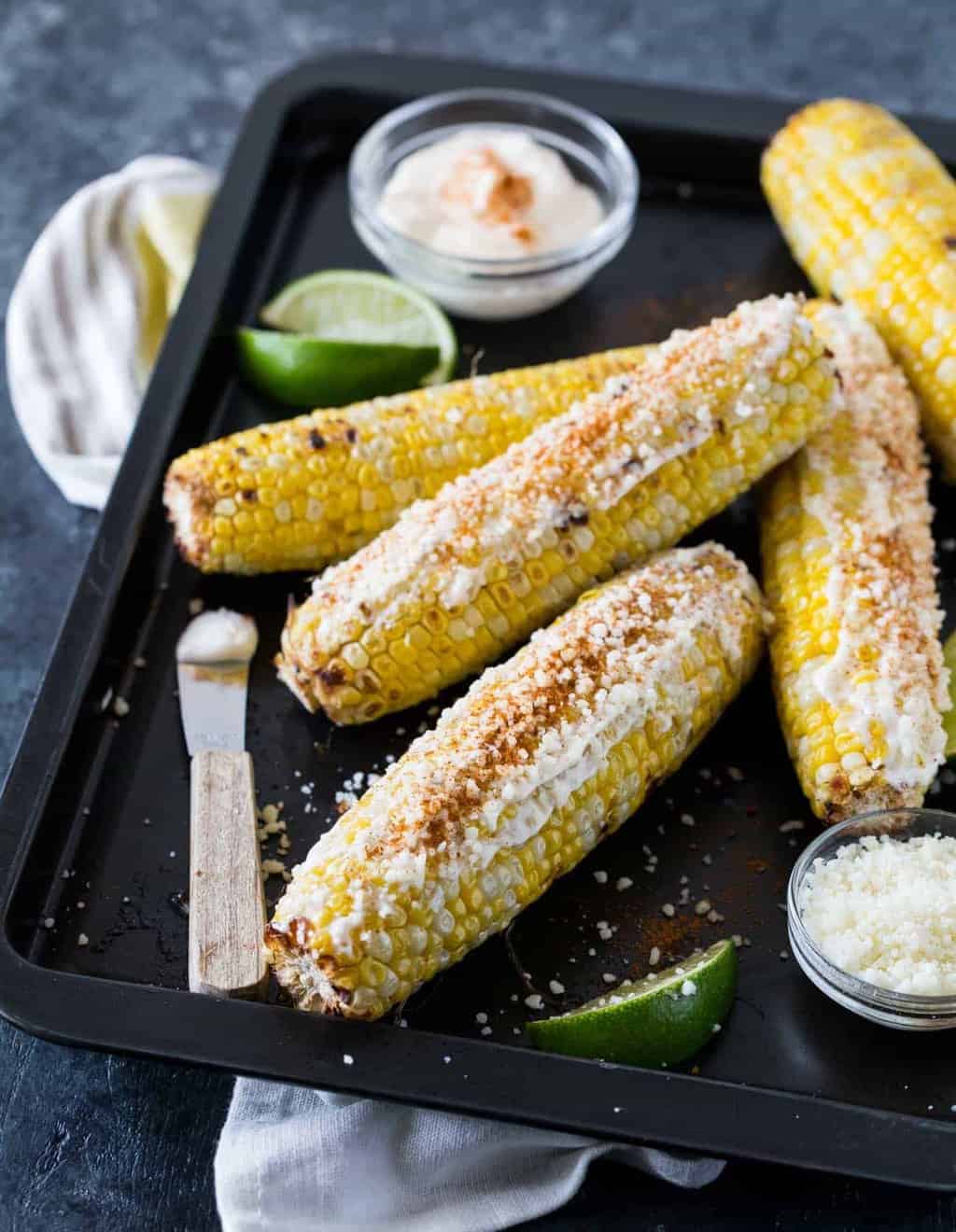 Mexican grilled corn with mayonnaise, cotija cheese, cayenne pepper and lime wedges piled high on a sheet pan.