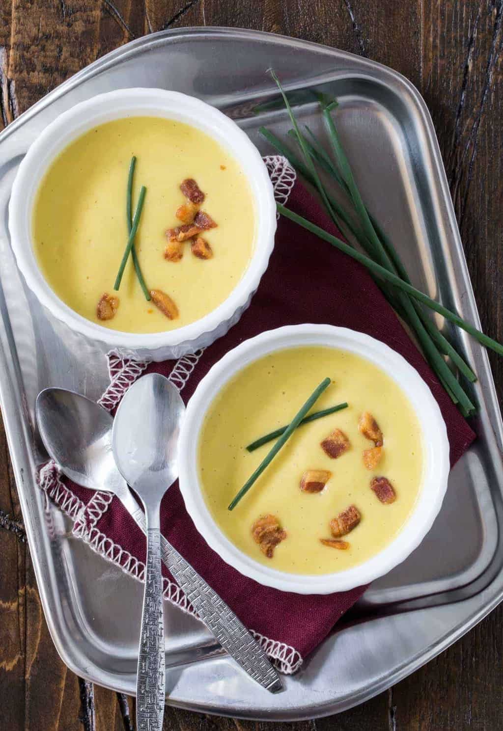 Two bowls of creamy Sweet Corn Soup on a silver tray garnished with crispy pancetta bits and chives.