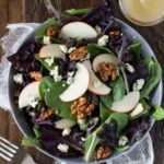 Silver bowl of Mixed Green Salad with Blue Cheese Crumbles, Apples and Candied Walnuts. Next to salad is a small glass bowl of salad dressing.