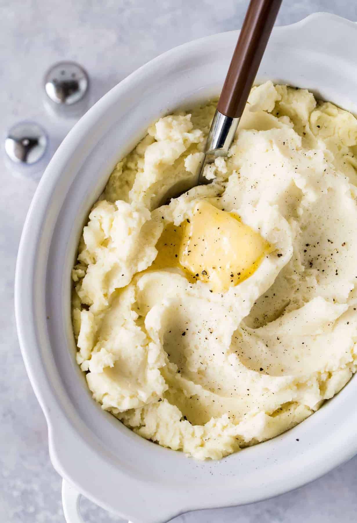 Slow cooker filled with mashed potatoes with melted butter on top. Salt and pepper shakers on side on white board.