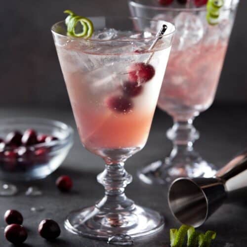 Cranberry gin cocktail in a footed glass filled with ice and garnished with cranberries and a lime twist.