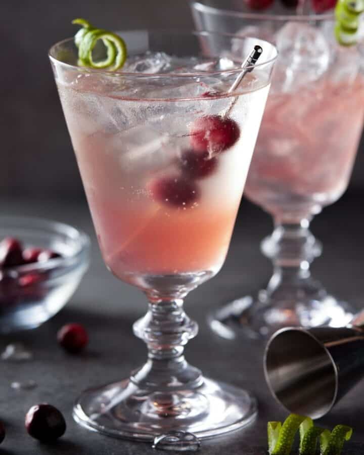 Cranberry gin cocktail in a footed glass filled with ice and garnished with cranberries and a lime twist.