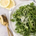 Large white bowl filled with Arugula Salad with Lemon Vinaigrette, with freshly shaved parmesan cheese and lemon wedges