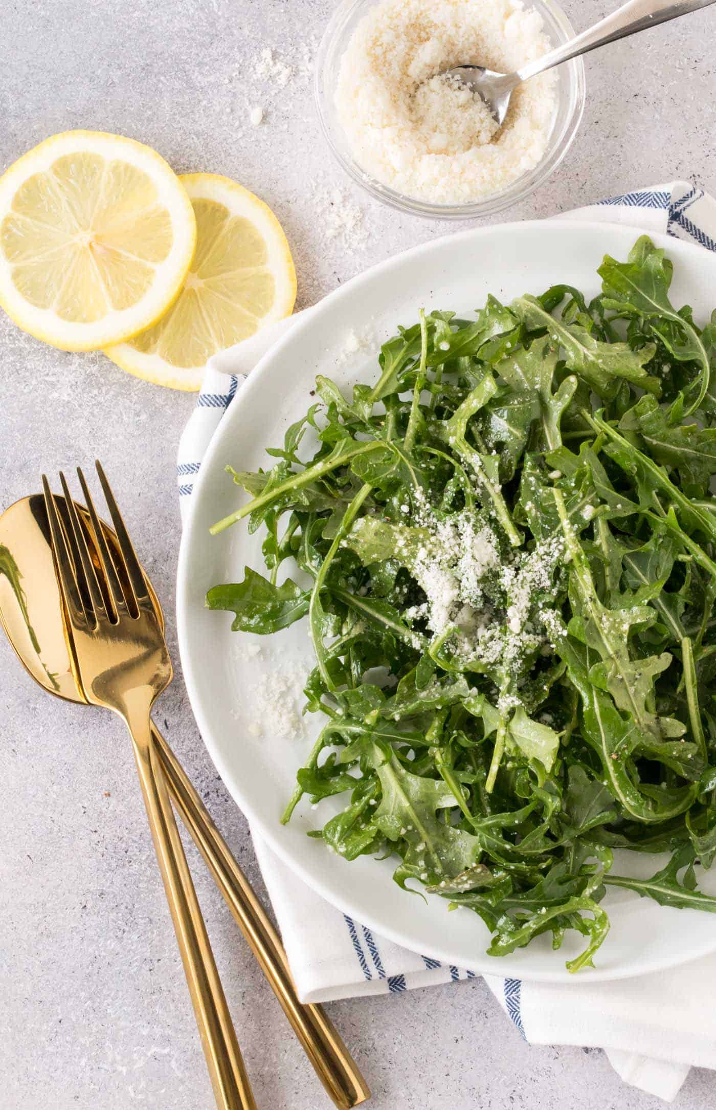 Large white bowl filled with Arugula Salad with freshly shaved parmesan cheese and lemon wedges