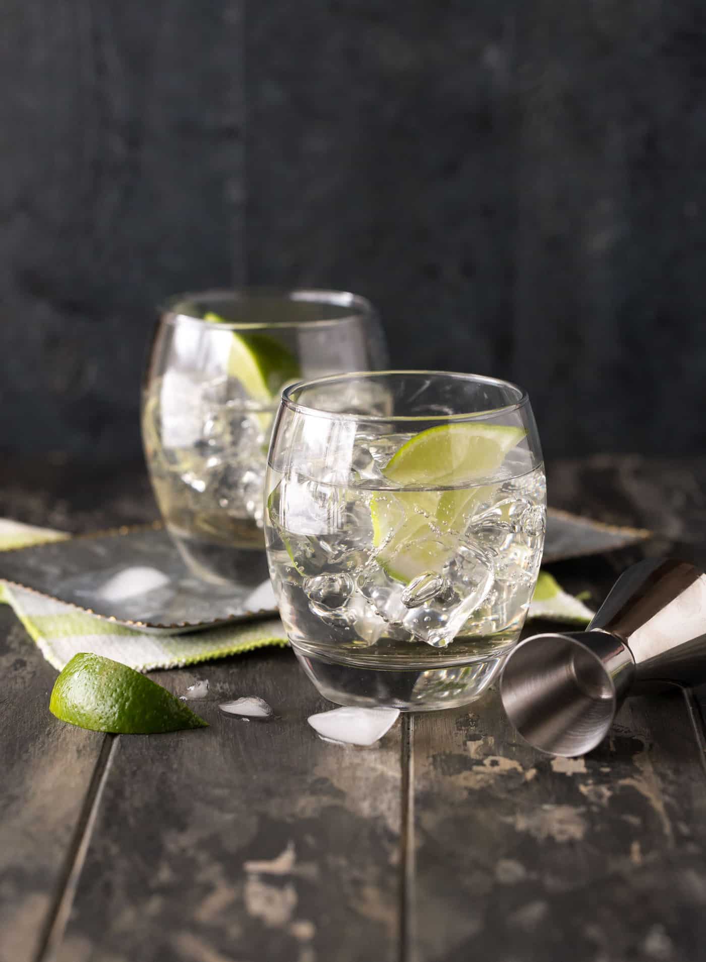 Deux verres lowball de cocktails Gin et Tonic à la fleur de sureau sur un tableau noir foncé, garnis de quartiers de lime et d'un jigger à cocktail.