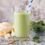 Glass of Easy Banana Spinach Protein Smoothie on a grey countertop and garnished with a blue and white striped straw.