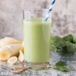 Glass of Easy Banana Spinach Protein Smoothie on a grey countertop and garnished with a blue and white striped straw.