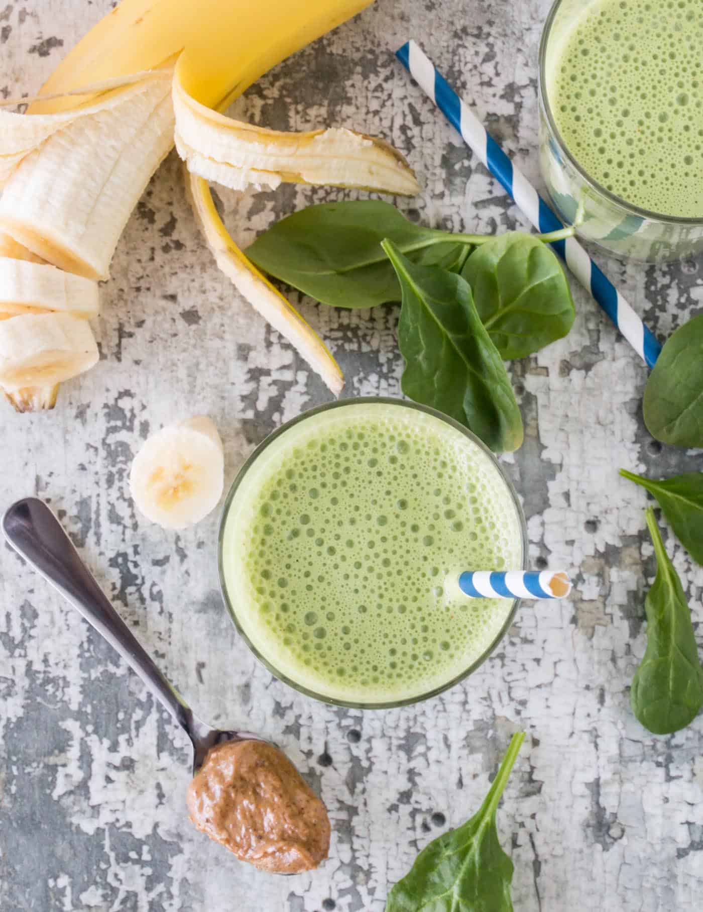 Two glasses of Smoothies on a gray countertop. Surrounded by blue and white striped straws, fresh banana slices, fresh spinach and a spoonful of almond butter.