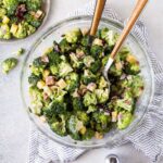 Glass bowl full of Easy Broccoli Salad with Bacon and Cheese garnished with large serving spoons and salt and pepper shakers.