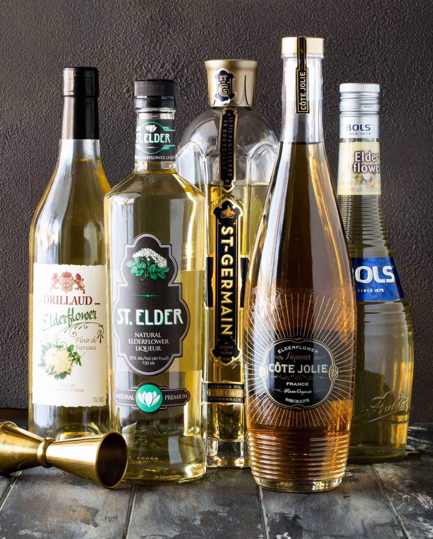 Bottles of different brands of elderflower liqueur with gold jigger.