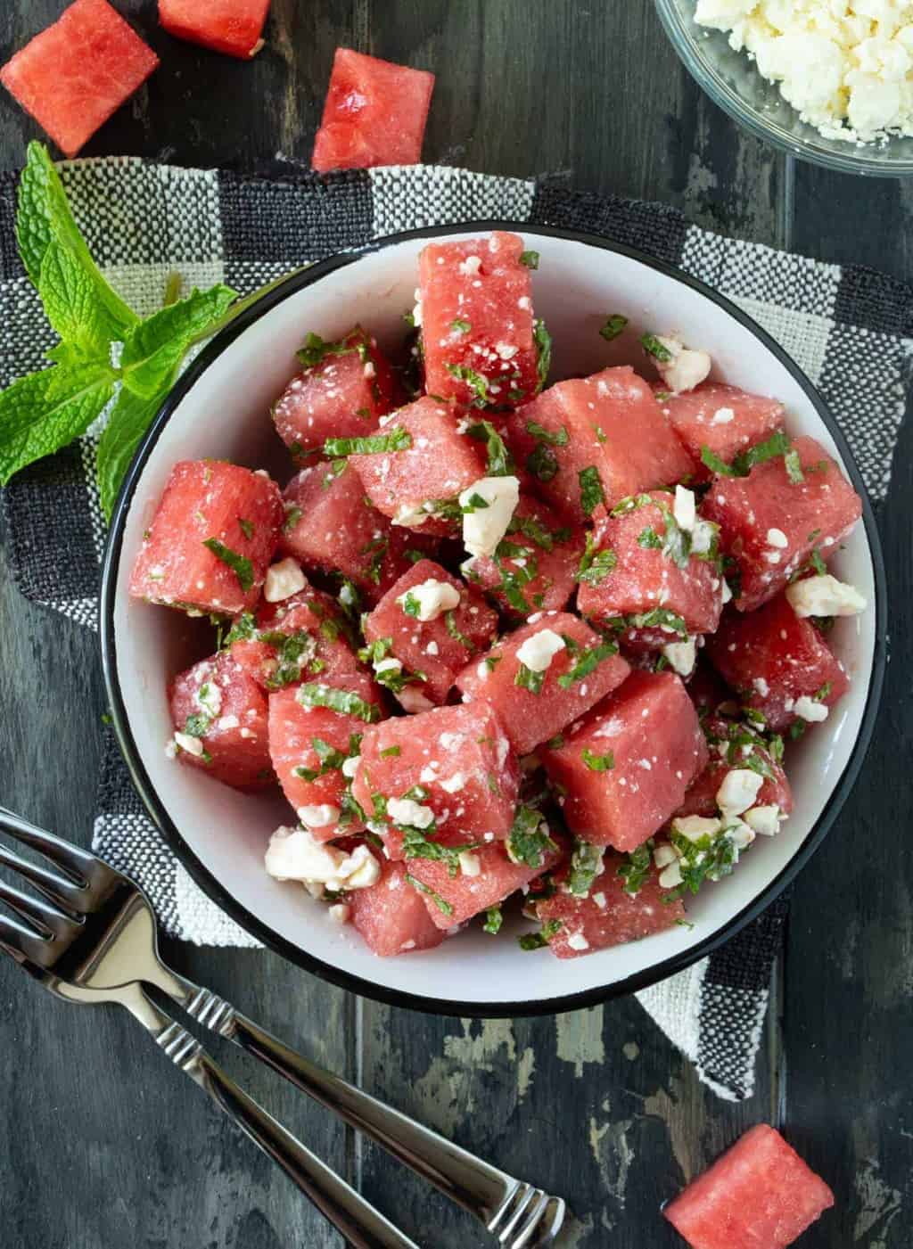 White bowl of Watermelon Feta Salad on a checkered napkin. Forks, watermelon cubes and feta cheese surround the bowl.