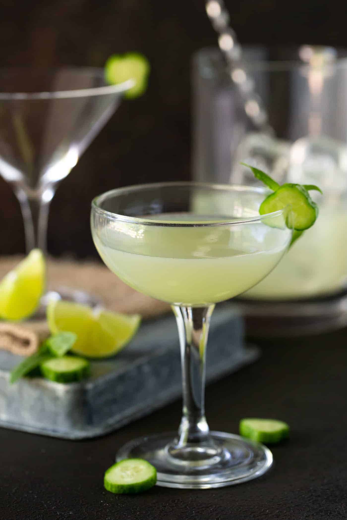 Cucumber Vodka Gimlet recipe in coupe glasses with a pitcher of the cocktail in the background.