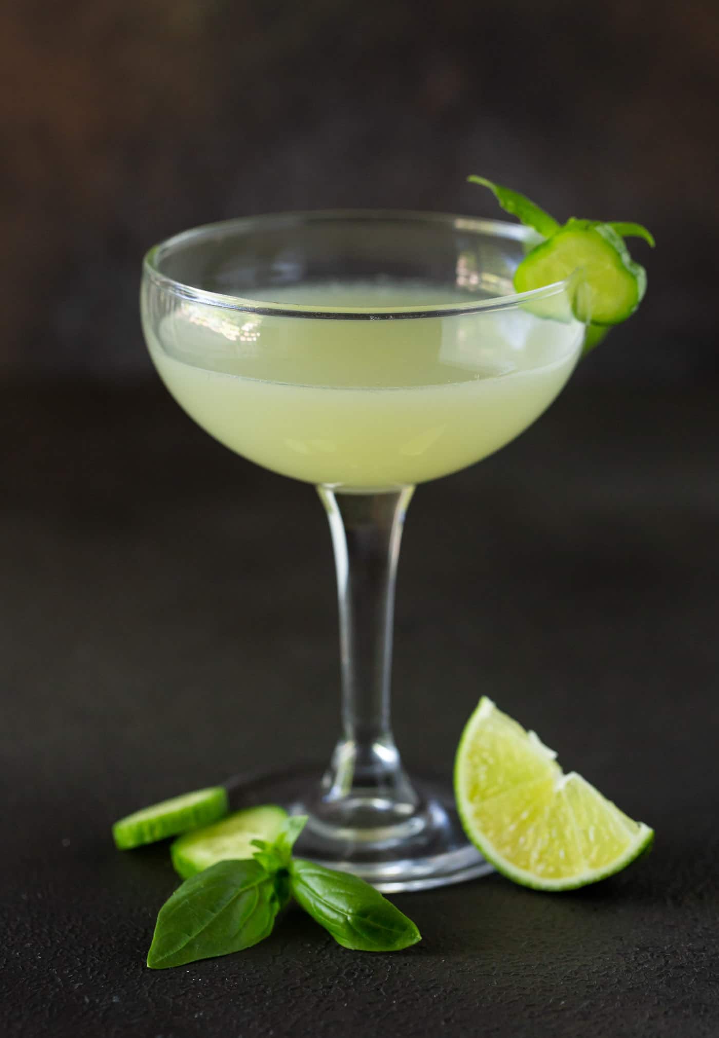 Gimlet in a coupe glass on a dark gray background. Garnished with lime wedges, fresh basil and cucumber slices.