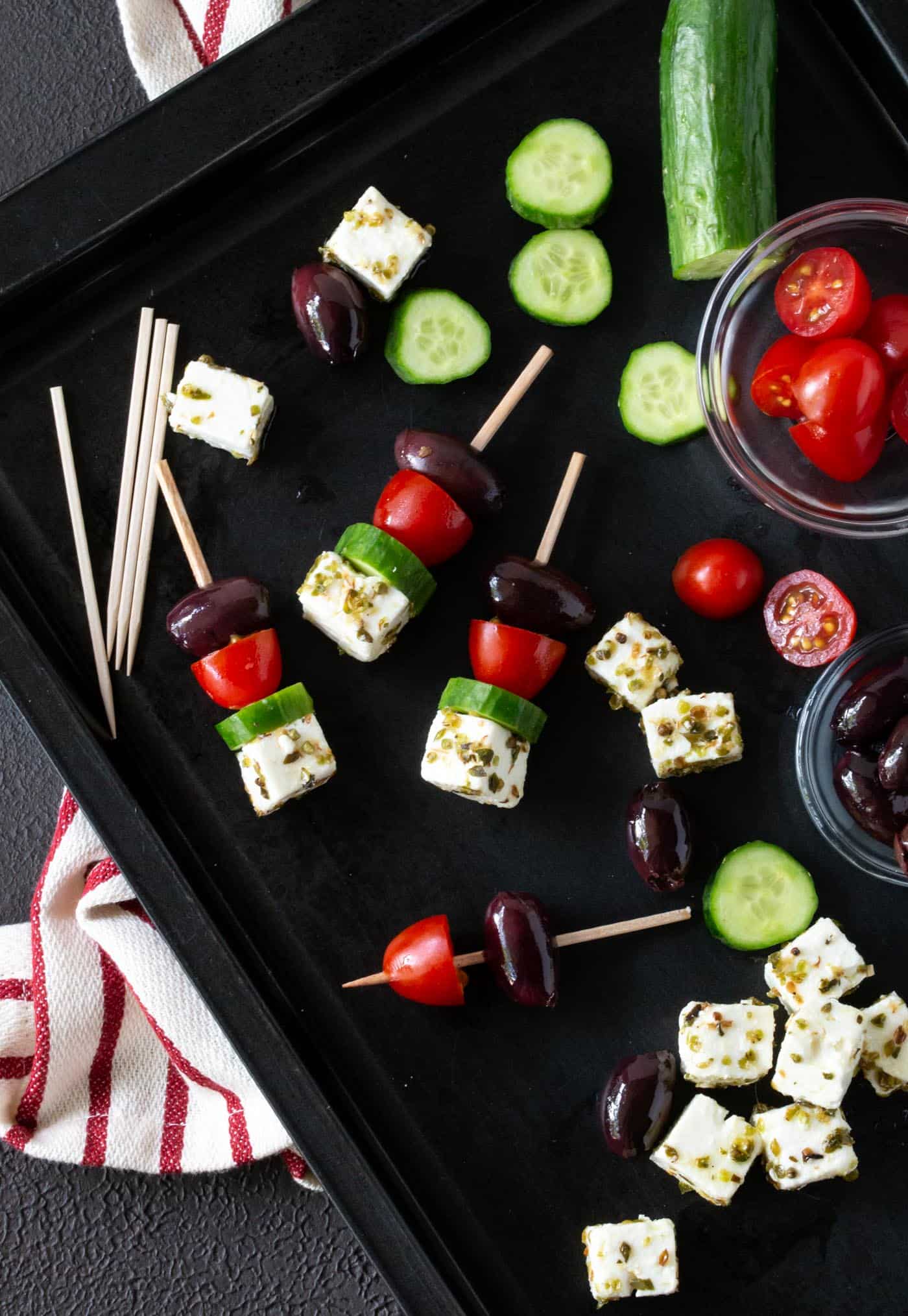 dark sheet pan filled with ingredients for Greek Salad Skewers including kalamata olives, feta cheese, grape tomatoes and cucumber chunks.