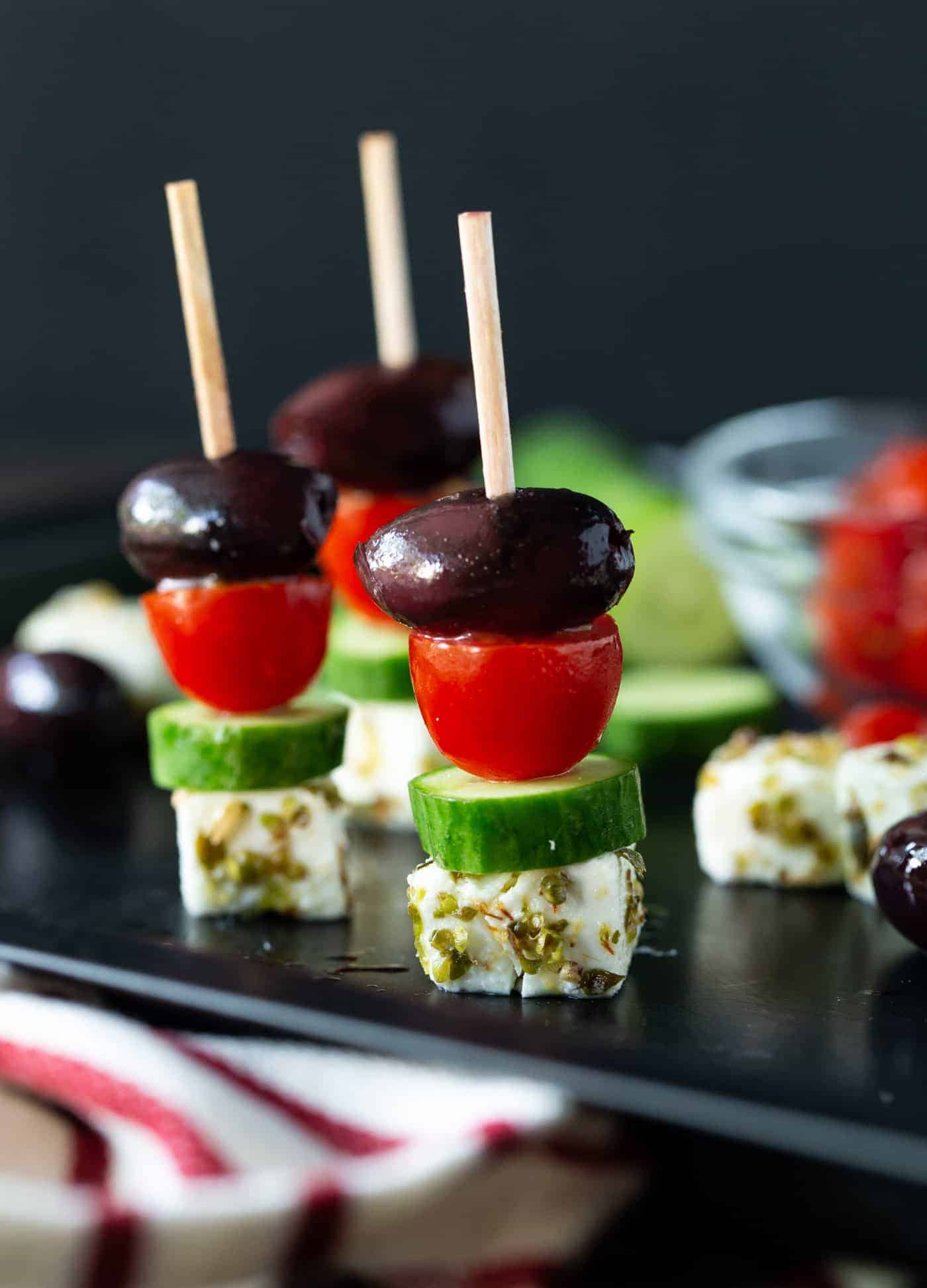 Three Greek Salad Skewers on dark platter with cubes of feta cheese, chunks of cucumber, kalamata olives and sliced tomatoes