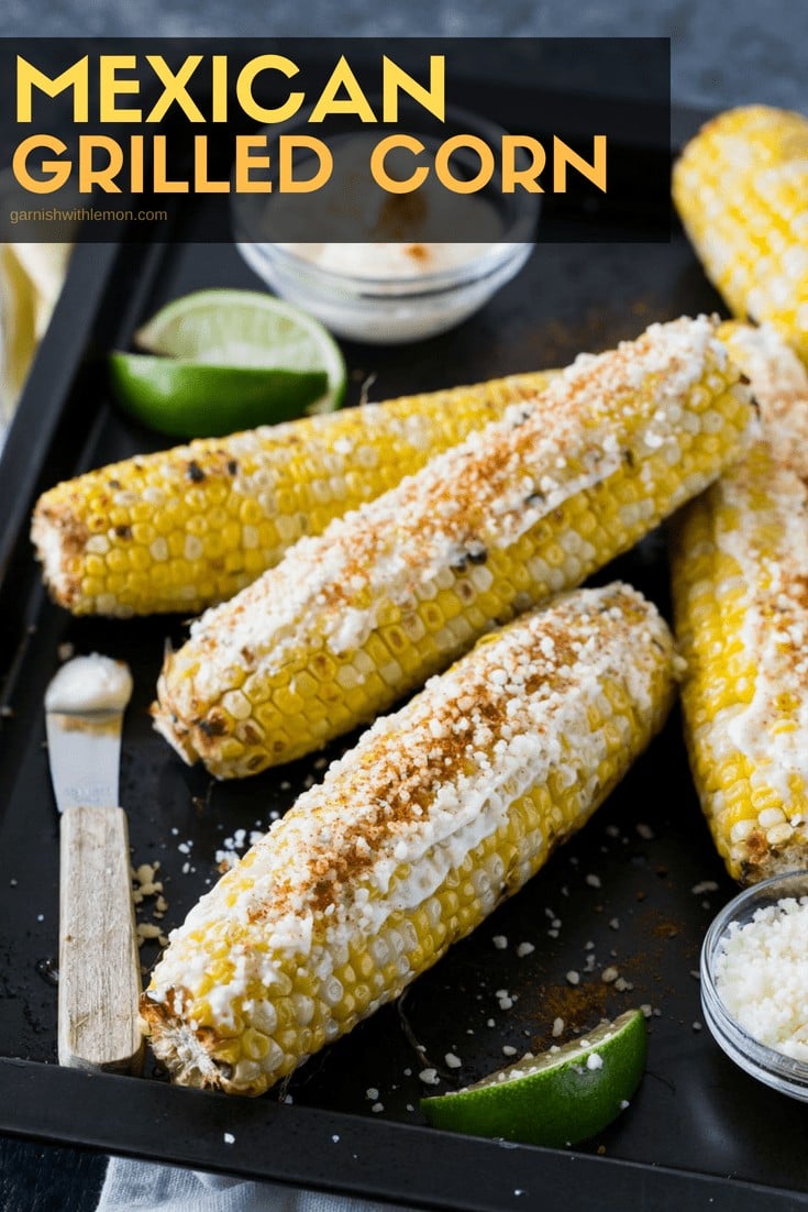 Sheet pan filled with Mexican Grilled Corn and garnished with lime wedges, cotija cheese and mayonnaise.