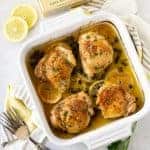 Chicken Thighs in white square baking dish with lemon slices and parsley for garnish.