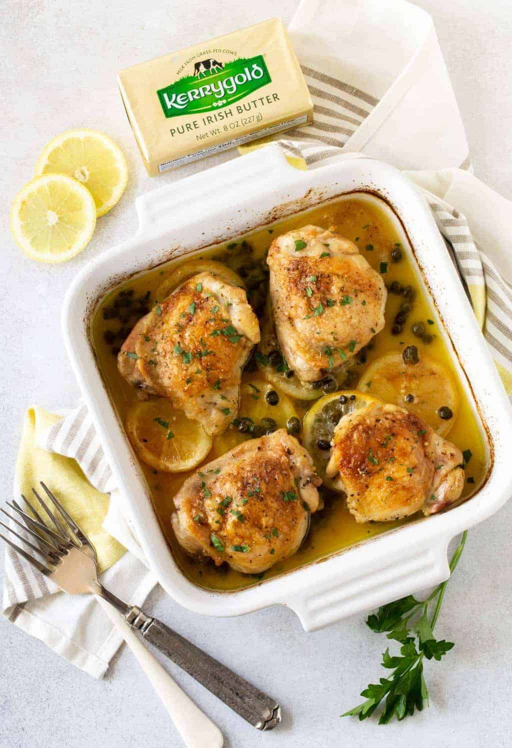  Chicken Thighs in white square baking dish with lemon slices and parsley for garnish.