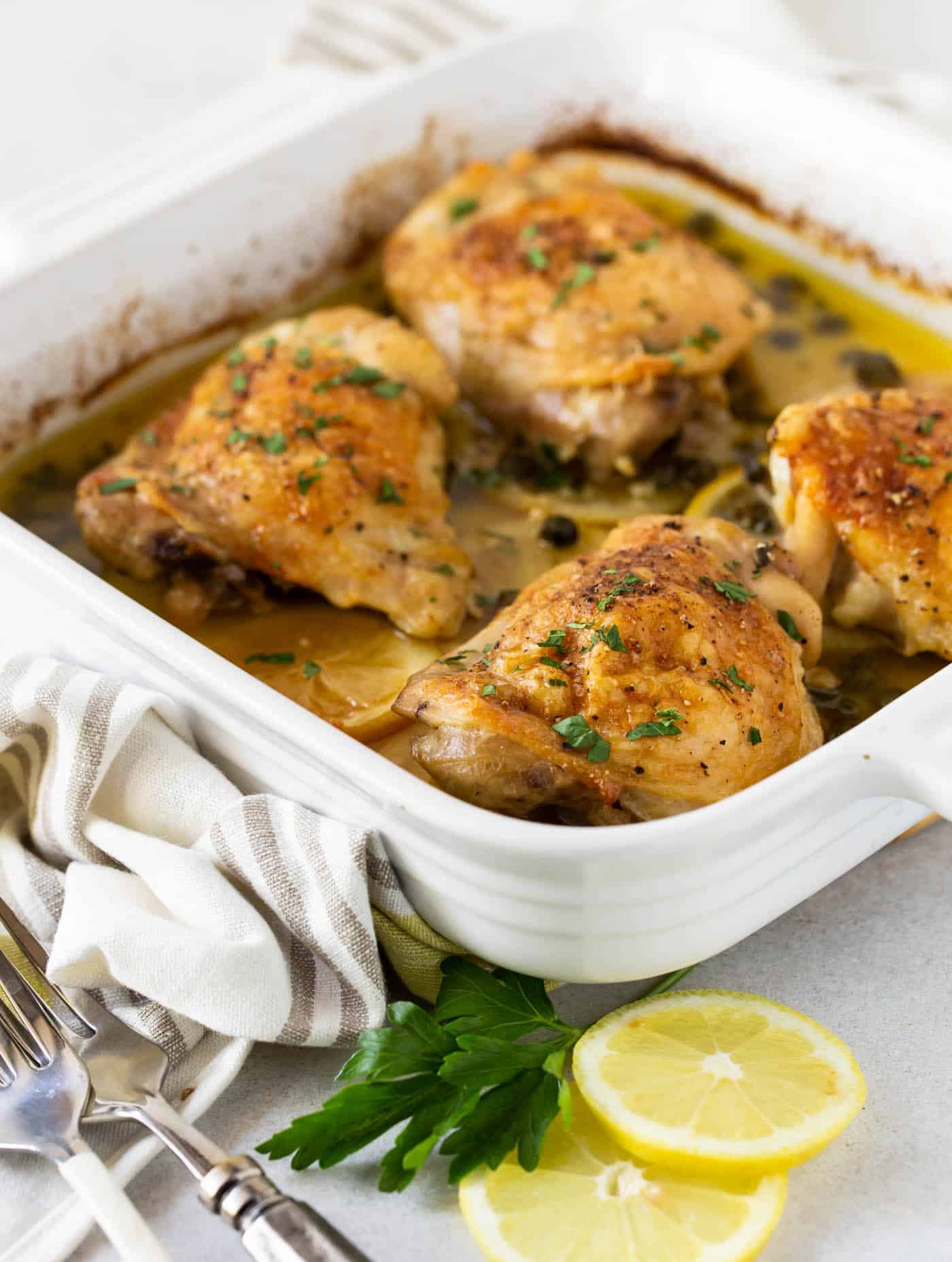 Chicken Thighs with Capers in a white baking dish with parsley and lemon slices for garnish.