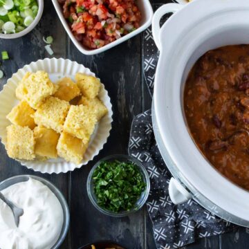 A white slow cooker filled with chili and surrounded by bowls of chili toppings like corn chips, pico de gallo, cornbread, cilantro, sour cream, green onions and shredded cheddar cheese.