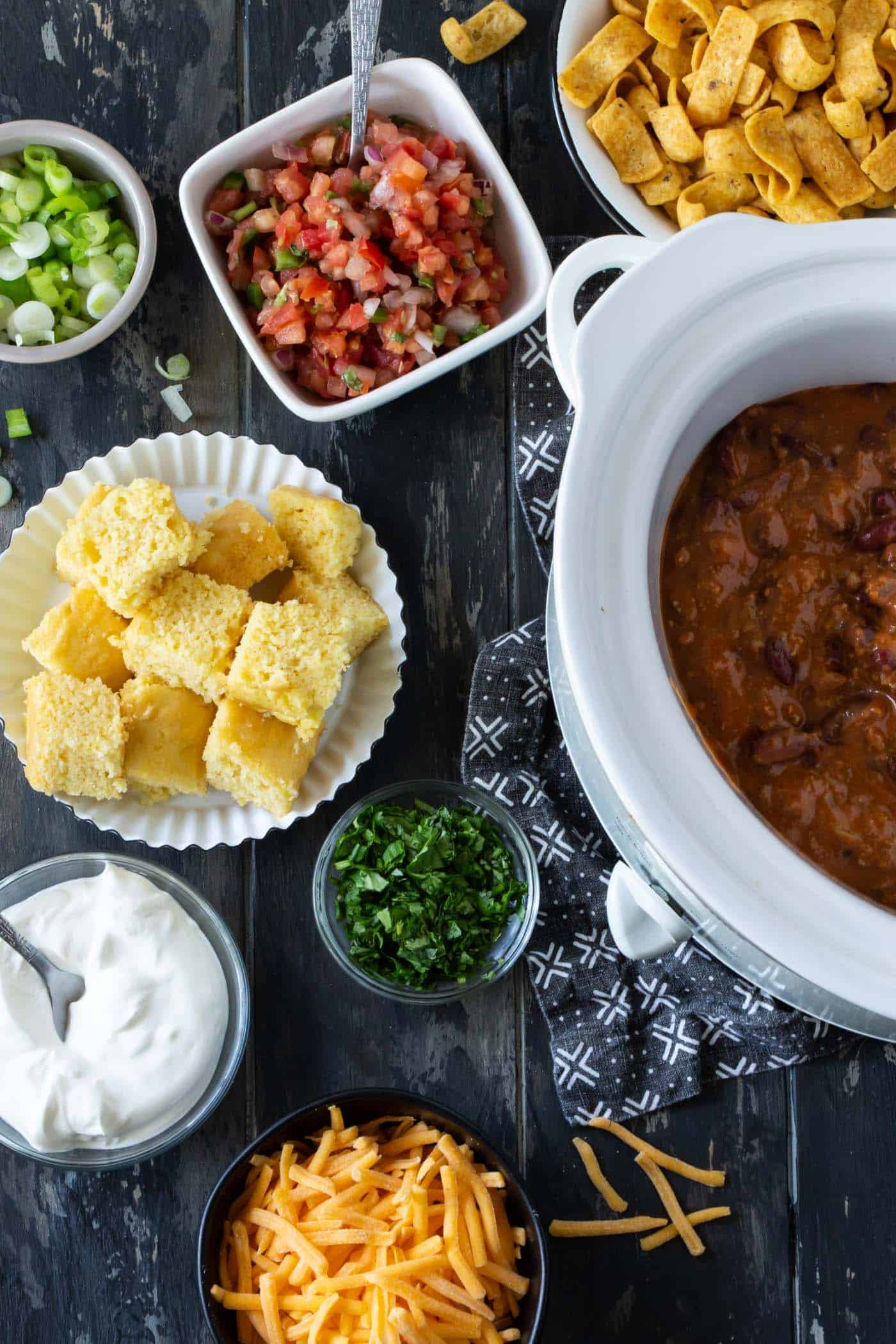 A white slow cooker filled with chili and surrounded by bowls of chili toppings like corn chips, pico de gallo, cornbread, cilantro, sour cream, green onions and shredded cheddar cheese.