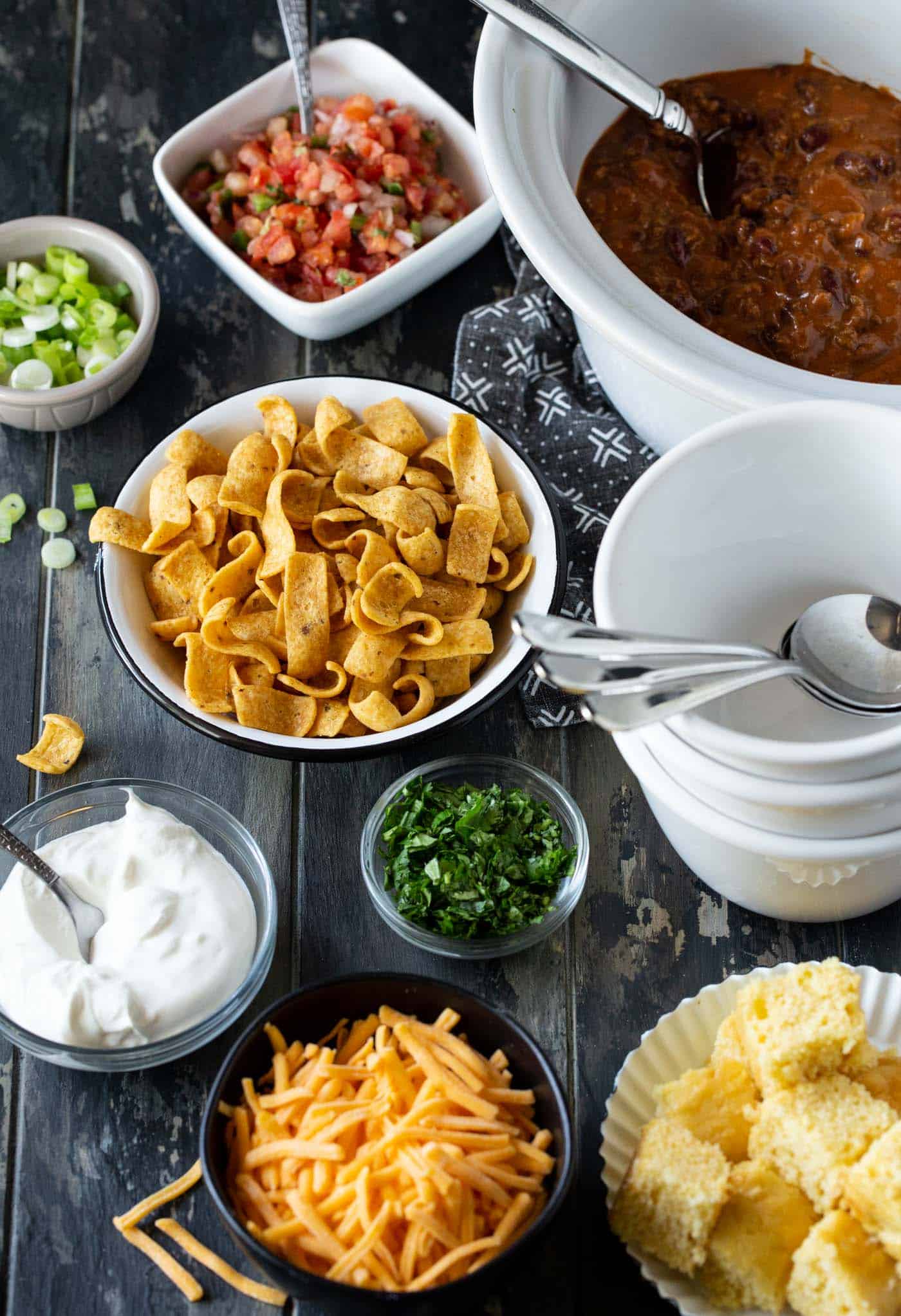 Slow cooker filled with chili. Surrounded by white chili bowls, spoons and chili toppings, including corn bread, fritos, pico de gallo, green onions, cilantro and sour cream.