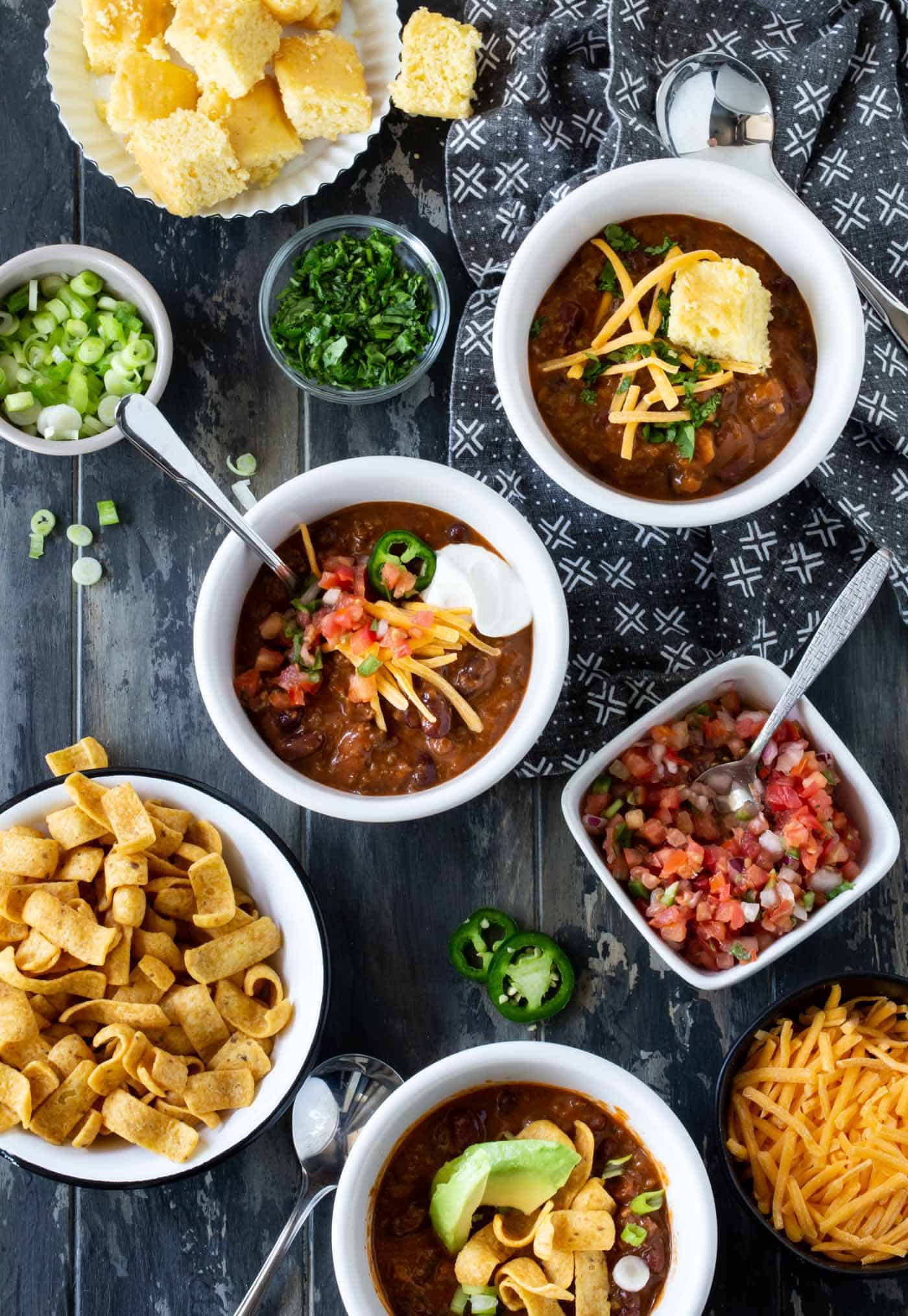 Three white bowls filled with chili and surrounded by bowls of chili toppings, including fritos, corn bread, green onions, pico de gallo, cilantro and shredded cheddar cheese.