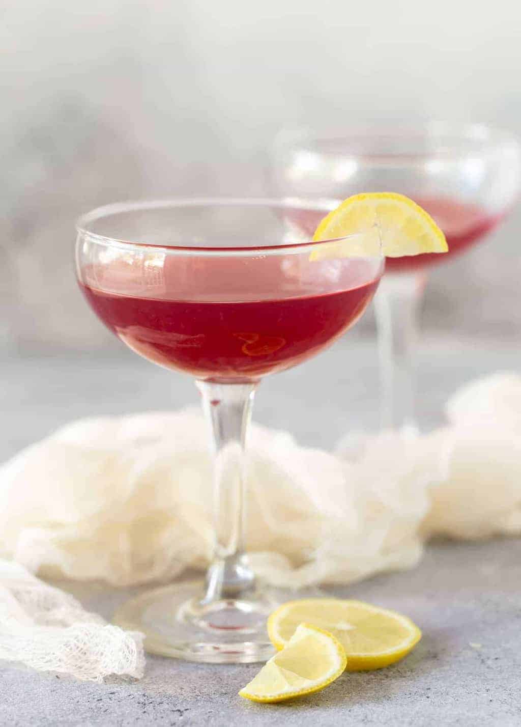 Two coupe glasses filled with Pomegranate Gin Cocktails on a grey board and garnished with small lemon slices.