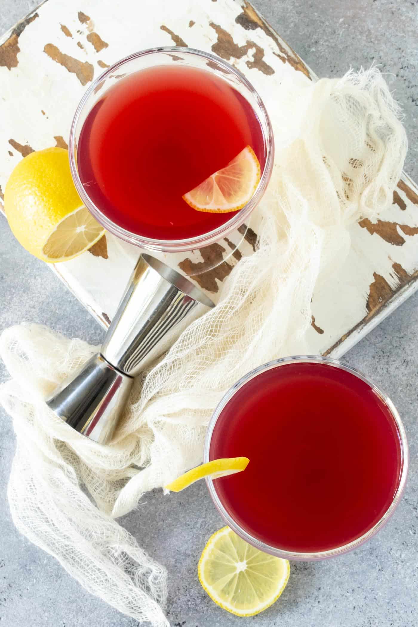 Top down shot of two coupe glasses filled with Pomegranate Gin Cocktails. Silver jigger and lemon slice garnishes surround glasses.