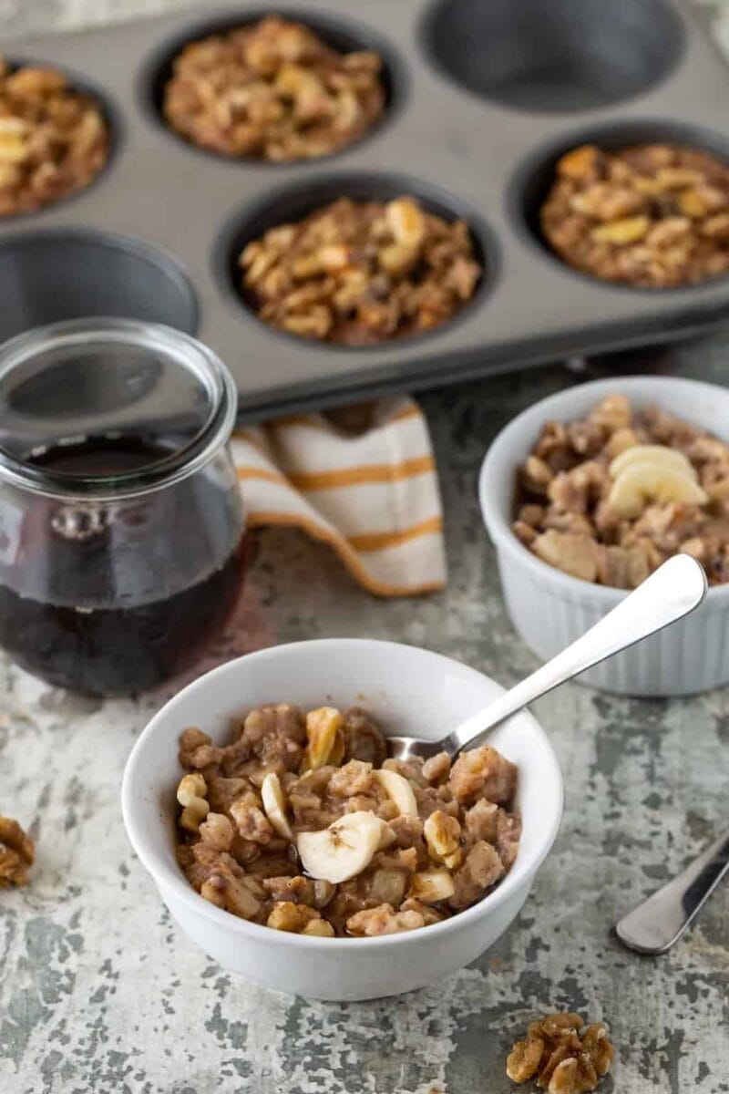 White bowls filled with Banana Nut Baked Oatmeal Cups. Muffin pan of oatmeal cups in the background. Garnished with maple syrup, walnuts and fresh banana slices.