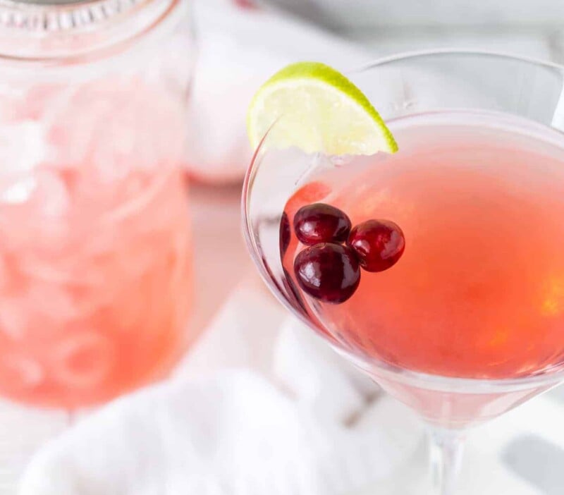 Martini in tall glass on a white background with fresh cranberries and lime wedges for garnish.