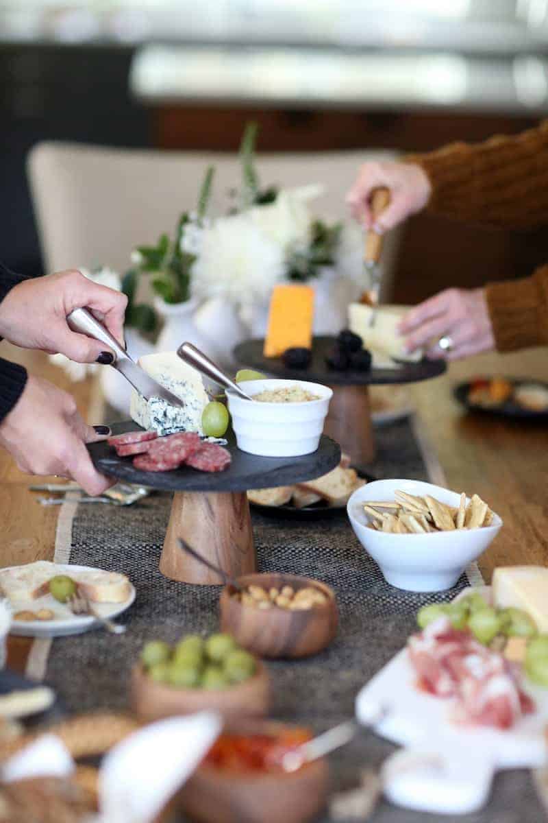 Hands cutting cheeses at a table with a plates of food with Garnish and Lemon.