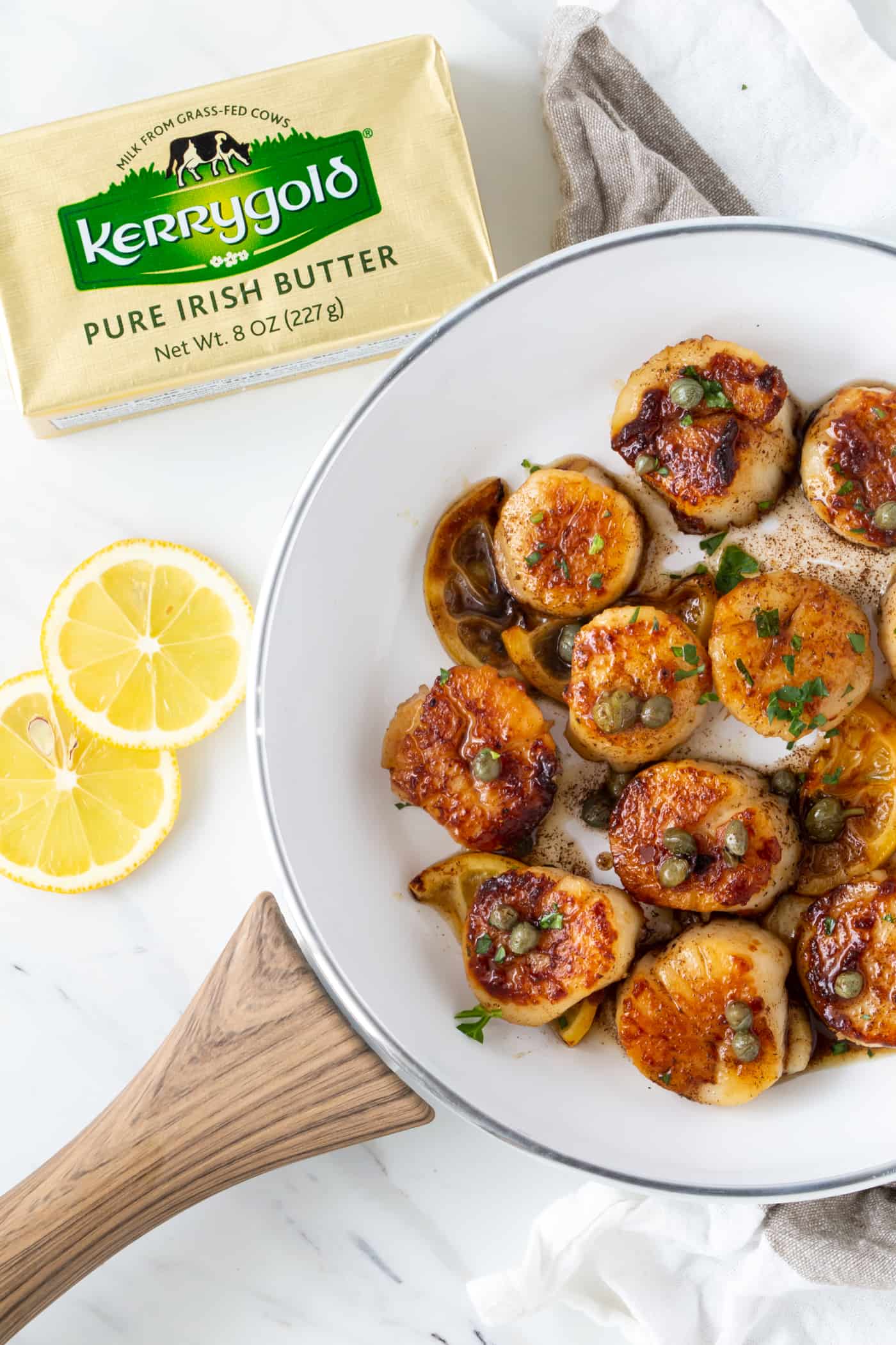  Scallops with brown butter lemon caper sauce in a white bowl with lemon slices and fresh parsley for garnish.