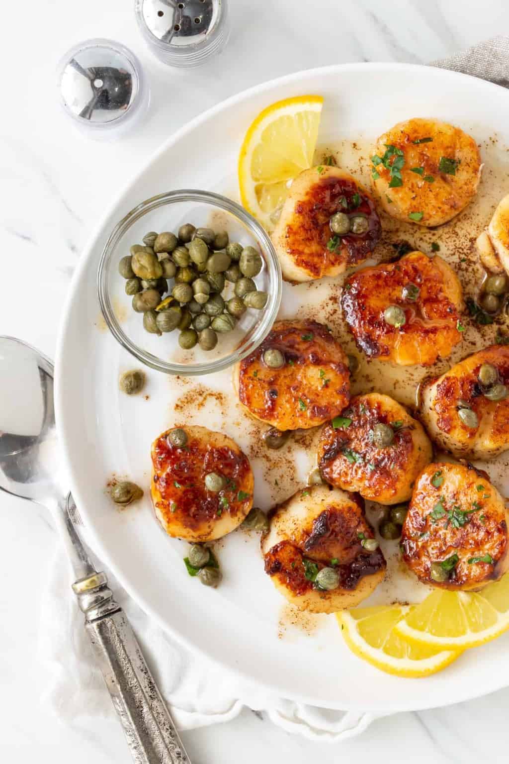  seared scallops in a white bowl with capers and lemon slices for garnish.