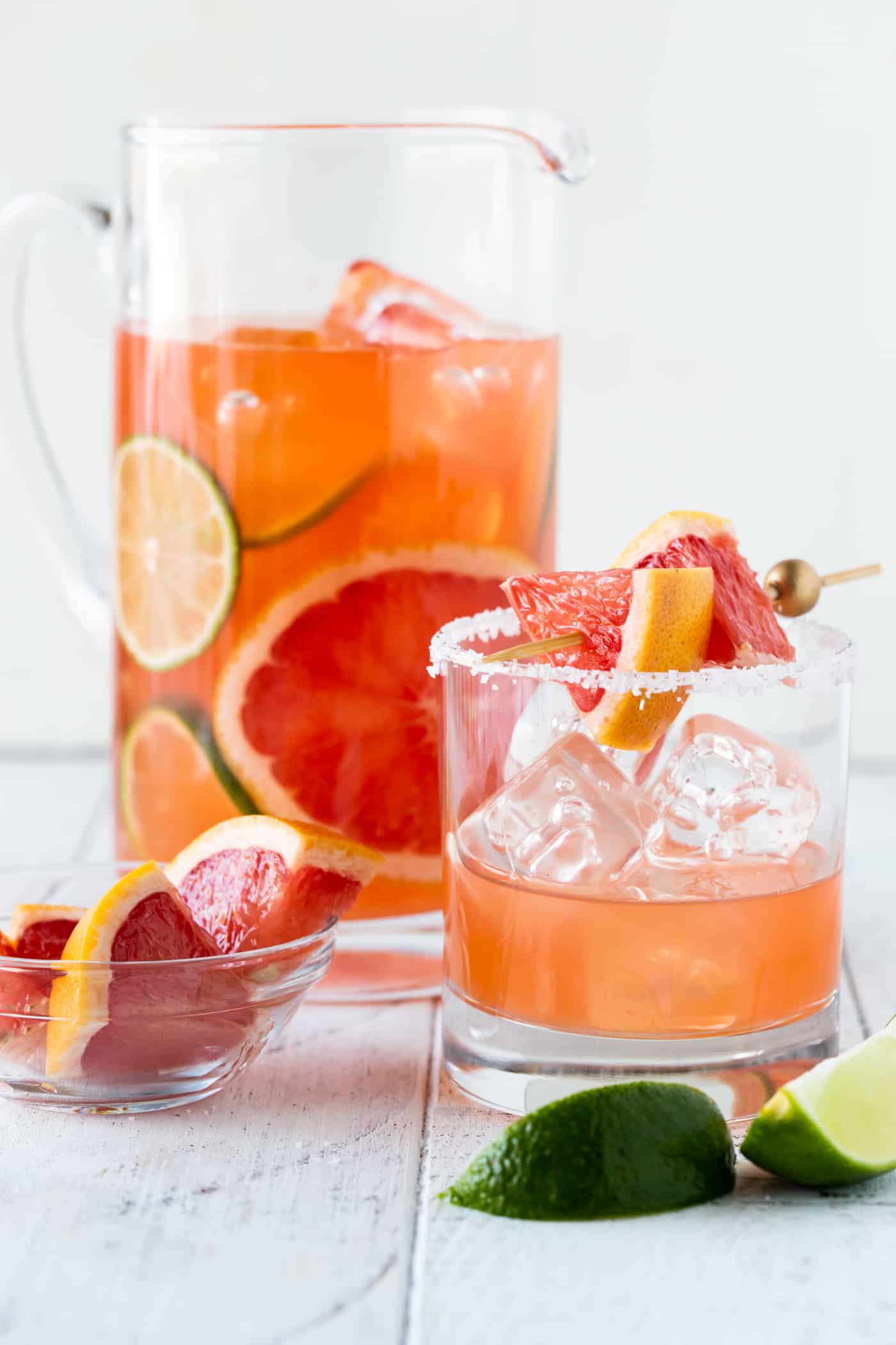 Grapefruit Aperol Cocktails in a low ball salt rimmed glass with grapefruit. pitcher of grapefruit aperol cocktails on white background.