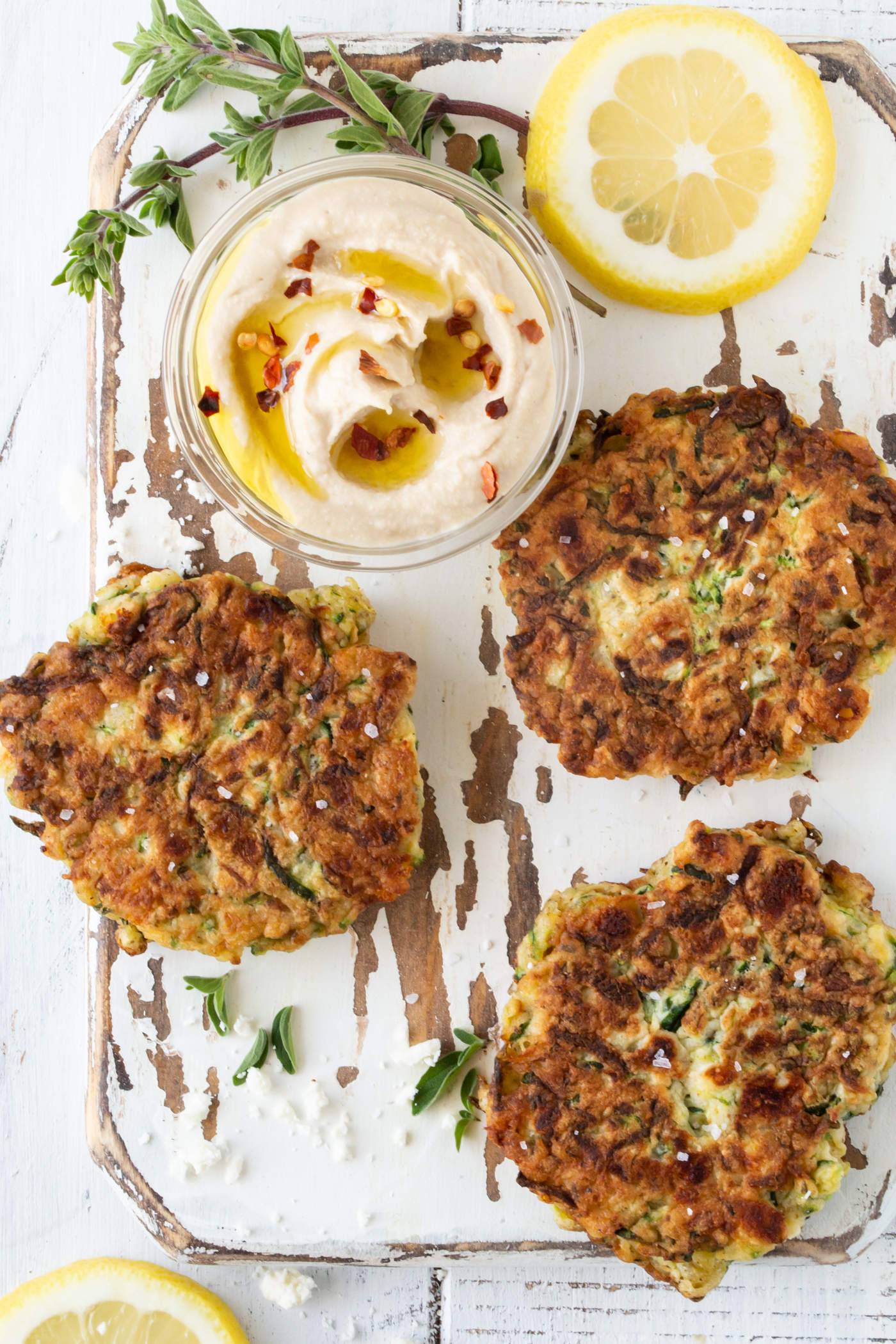 Three zucchini feta fritters on a white board. Garnished with coarse sea salt, hummus, fresh oregano and lemon slices.