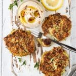 Zucchini Feta Fritters on a white board with a small bowl of hummus. Garnished with fresh oregano and lemon slices.